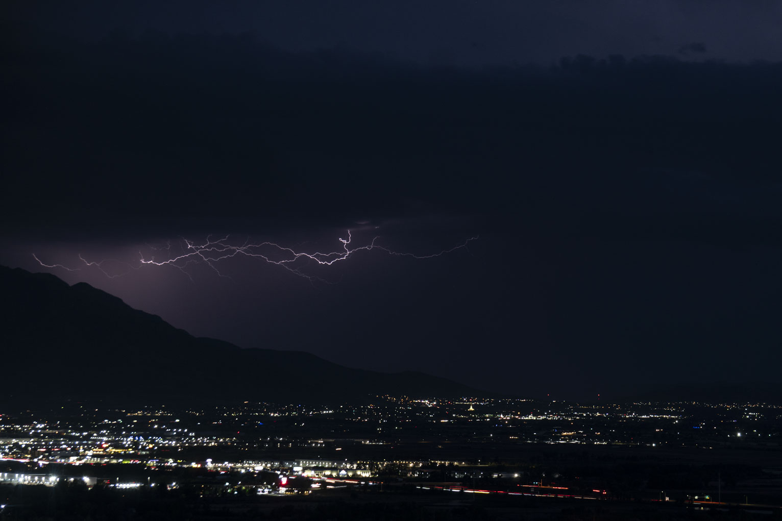 Lightning creeping on the underside of the clouds lights the surrounding mist purple above a town near the mountain